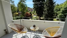 Balcony with two modern chairs, a small round glass table, and a decorative vase, overlooking a lush green area with trees.