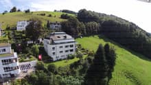 Aerial view of a modern white building with a flat roof and large windows, surrounded by lush green hills and trees. The building has a Swiss flag displayed in front.