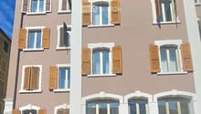 A three-story residential building with beige and brown facade, multiple windows with wooden shutters, and a balcony on the top floor.