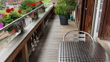 Wooden balcony with metal table and chair, potted plants, and scenic countryside view.
