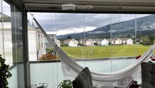 Balcony with large windows offering a scenic view of a green field, mountains, and buildings. Features a hanging chair, a small table, potted plants, and a hammock.