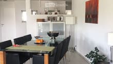 Modern dining area with a glass dining table, black chairs, a floor lamp, a cabinet, and artwork on the wall.