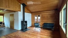 Spacious living room with wooden floors, exposed wooden ceiling beams, and a central fireplace. Features a dark green sofa and matching armchair, with large windows and a door leading outside.