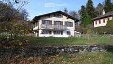 Two-story house with a balcony, wooden shutters, and a stone retaining wall in front.