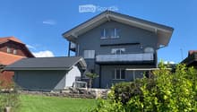 Two-story modern house with grey exterior, white trim, multiple windows, and a balcony on the upper level. The house features a well-maintained lawn and surrounding greenery.