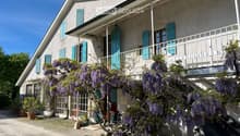 Building. Two-story house with grey stone facade, teal shutters, and a covered balcony adorned with blooming wisteria. The entrance features a bicycle and potted plants.