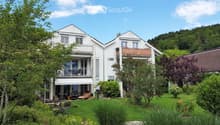 A two-story white house with balconies, surrounded by a well-maintained garden with outdoor furniture and greenery.
