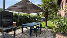 Garden with a table, chairs, a hammock, and a large umbrella, surrounded by greenery and a wooden stove.