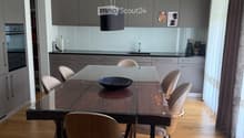 Modern kitchen with grey cabinets, a glass dining table, six chairs, a bowl on the table, and a hanging pendant light. The kitchen features a sleek design with a wooden floor and large windows with curtains.