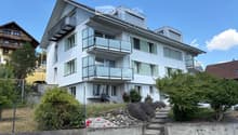Modern two-story house with white exterior, multiple balconies, large windows, and landscaped garden with steps leading up to the entrance.