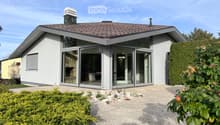 Modern single-story house with white exterior, large glass sliding doors, and a tiled roof. The house features a raised ground floor and is surrounded by neatly trimmed hedges and gravel pathways.