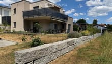 Modern two-story house with a balcony, patio seating area, and garden with stone wall and plants.