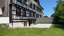 Traditional half-timbered house with white plaster walls, wooden beams, and multiple windows. The house has a sloped roof and is surrounded by a well-maintained lawn and a stone wall.