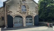 Stone building with arched doors and windows, surrounded by greenery and a paved area.