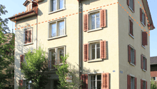 A multi-story residential house with light yellow walls, red shutters, and multiple windows. Features include a balcony on the second floor, a small garden area, and a staircase leading to the entrance.