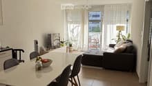 Open-plan living and dining area with a sofa, ottoman, dining table, chairs, and a TV. Large windows with curtains lead to a balcony with outdoor furniture.
