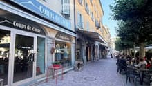 A charming pedestrian street lined with colorful buildings, featuring a clothing store named "Coups de Coeur" on the left and outdoor seating for a café on the right. The street is paved with cobblestones and has trees lining the far side.