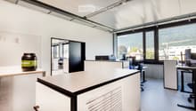 Modern office space with white walls, large windows, desks, chairs, and a large screen. A large container is placed on a table near the window.