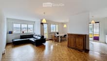 Open-plan living and dining area with wooden flooring, large windows offering a scenic view, a black leather sofa, a dining table with chairs, and a wooden cabinet.