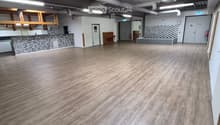 Empty room with wooden flooring, white walls, and exposed industrial-style ceiling with pipes and lighting. Features include a hexagonal-patterned counter, a small seating area, and a doorway leading to another room.