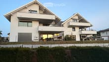 Modern two-story house with white exterior, large windows, balconies, and a garage. The house is situated on a raised ground floor with a well-maintained lawn and hedge in front.