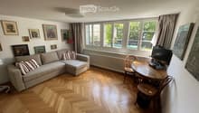Living room with light wooden herringbone flooring, large windows with curtains, a cream sectional sofa with striped cushions, a wooden coffee table with a TV, and several framed artworks on the walls.