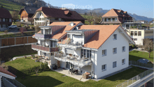 A modern two-story house with a red-tiled roof, white exterior, and multiple balconies. The house is surrounded by a well-maintained lawn and features outdoor seating areas and a patio.
