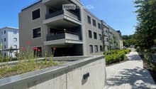 multistory building, balconies, gray walls, bricked pathway, shrubs, parking area in front