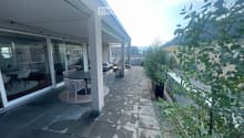 Modern balcony with tiled floor, outdoor furniture including a table and chairs, large glass sliding doors leading to indoor space, and lush green plants.
