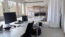 Modern office space with large windows, white walls, and a grey carpeted floor. A white desk holds a computer monitor, keyboard, mouse, and office supplies. An office chair is positioned in front of the desk. A black curtain is on the right side, and shel