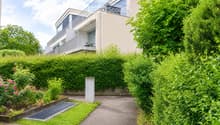Modern residential building with raised ground floor, surrounded by lush greenery and a well-maintained garden path.