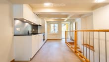 Modern hallway with tiled floor, white walls, built-in kitchen area on the left, staircase with wooden railing leading upwards, and a window at the end.