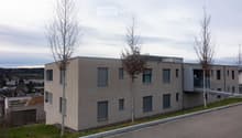 Modern two-story building with beige facade, multiple windows, and a balcony on the upper level. The building is surrounded by a few trees and a paved road.