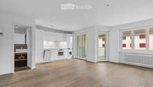 Completely empty room with wooden flooring, white walls, and modern white kitchen cabinets visible in the background. No furniture or equipment present.