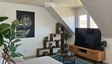 Modern living room with a white sofa, coffee table, TV stand, shelves, and potted plants. The room features a slanted wooden ceiling and large windows.