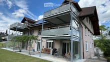 Modern two-story house with a large balcony and patio area, featuring outdoor furniture and a well-maintained lawn.