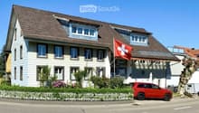 Two-story house with a brown tiled roof, white siding, multiple windows, and a Swiss flag. A red SUV is parked in front, and there is a small garden with a white picket fence.