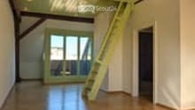 Bright, open loft space with exposed wooden beams, polished wooden floor, large windows, and a small staircase leading to an upper level.