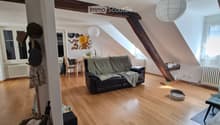 Living room with wooden flooring, slanted ceiling, exposed wooden beams, and large windows. Features a black leather sofa with a green blanket, a small table with chairs, a hanging paper lantern, a speaker, and a cat bowl on the floor.