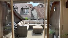 A modern balcony with a grey wicker sofa, ottoman, small table, and potted plants, overlooking a cityscape with traditional rooftops.