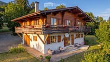A two-story wooden house with a large balcony, surrounded by greenery and a well-maintained lawn. The house features a mix of wooden and white exterior walls, multiple windows with shutters, and a chimney. Outdoor seating includes a sofa, chairs, and a sm