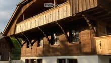 Two-story wooden house with a balcony, large windows, and decorative wooden trim. The lower level has a white plaster finish and potted plants near the entrance.