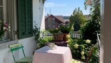 A cozy balcony with a small table and chairs, surrounded by lush greenery and flowers, with a view of the garden and neighboring houses.
