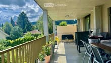Balcony with a sofa, small table, chairs, potted plants, grill, and storage bin, overlooking a green landscape.