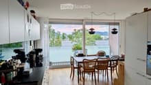 Open-plan kitchen and dining area with modern white cabinets, a black countertop, and a dining table with six chairs. The space features large windows with a view of greenery and a balcony area.