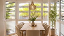 Bright dining area with a wooden table, six chairs, a vase with greenery, and a candle on the table. Large windows with blinds offer natural light and a view of greenery outside.