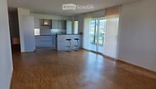 Empty room with wooden flooring, white walls, and large windows with blinds. Kitchen area visible in the background with cabinets and a countertop.