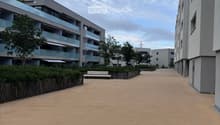 A wide, sandy pathway between modern apartment buildings with balconies, surrounded by greenery and a wooden bench.