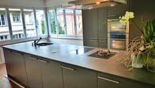 Modern kitchen with grey cabinets, a central island with a sink and stove, and large windows overlooking the street.