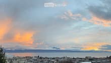 Panoramic cityscape view at sunset, featuring a body of water, distant mountains, and urban buildings.